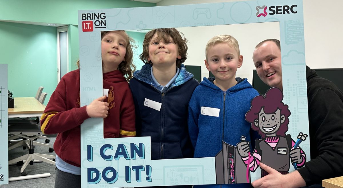 Three children smiling to camera with male lecturer, all behind Bring IT On prop frame with I can do I.T. at bottom.